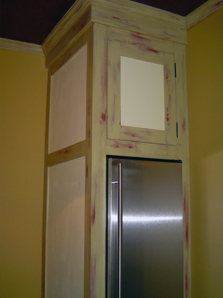 Pantry cabinetry painted with pale yellow-green and rubbed through to reveal red undercoat and oak wood grain relief.