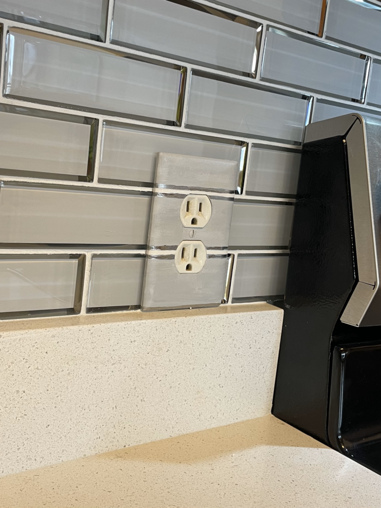Series of outlet cover plates painted to blend into grey and chrome glass backsplash.
